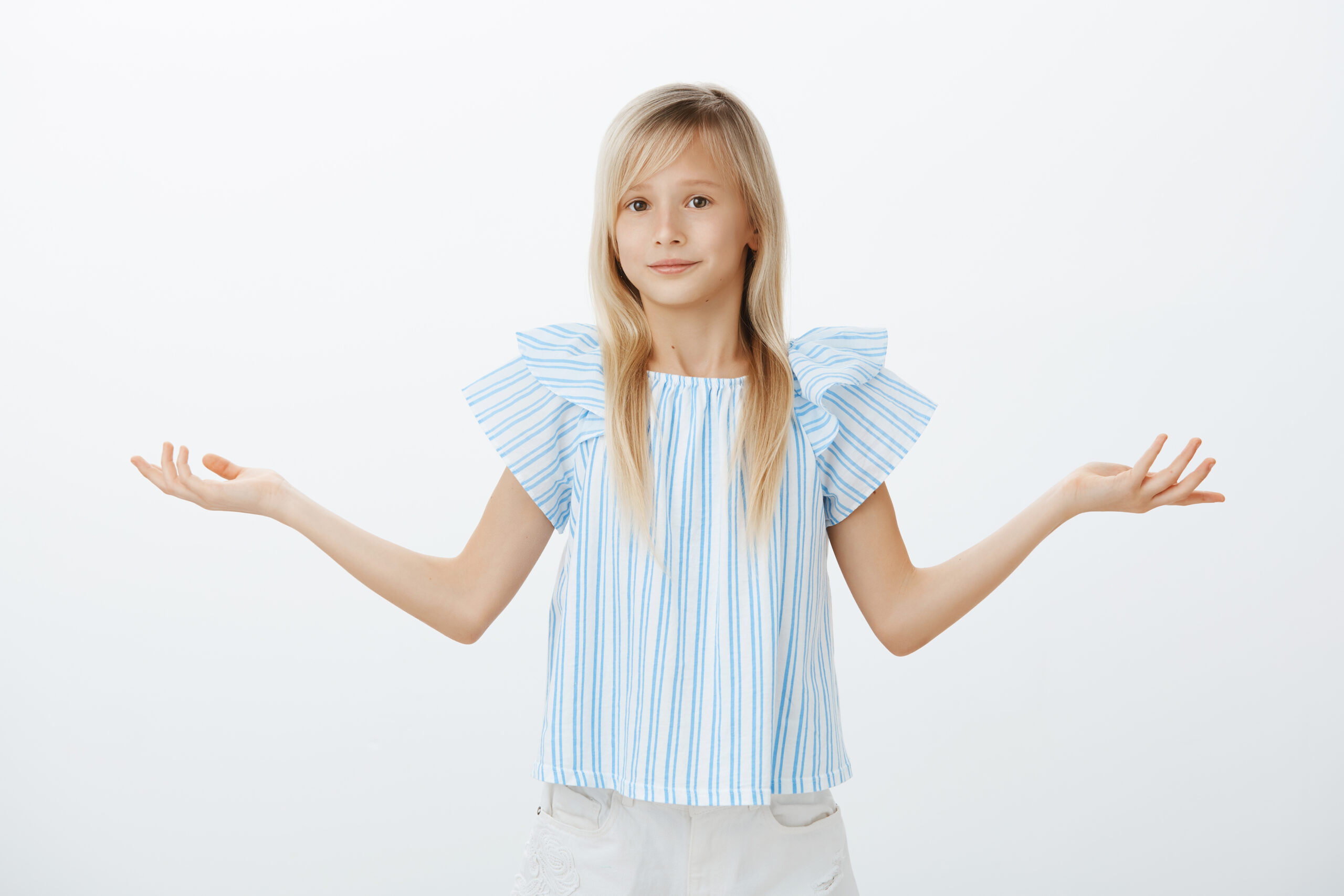 Portrait of intense unaware cute little girl with blond hair, shrugging with spread palms, smiling with awkward problematic expression, being unable to answer, standing clueless over gray background