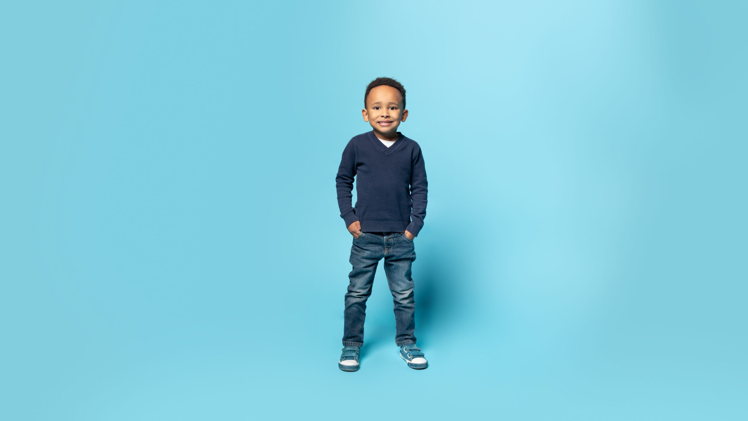 Happy childhood. Full length shot of black little boy holding hands in pockets, blue background, panorama, free space