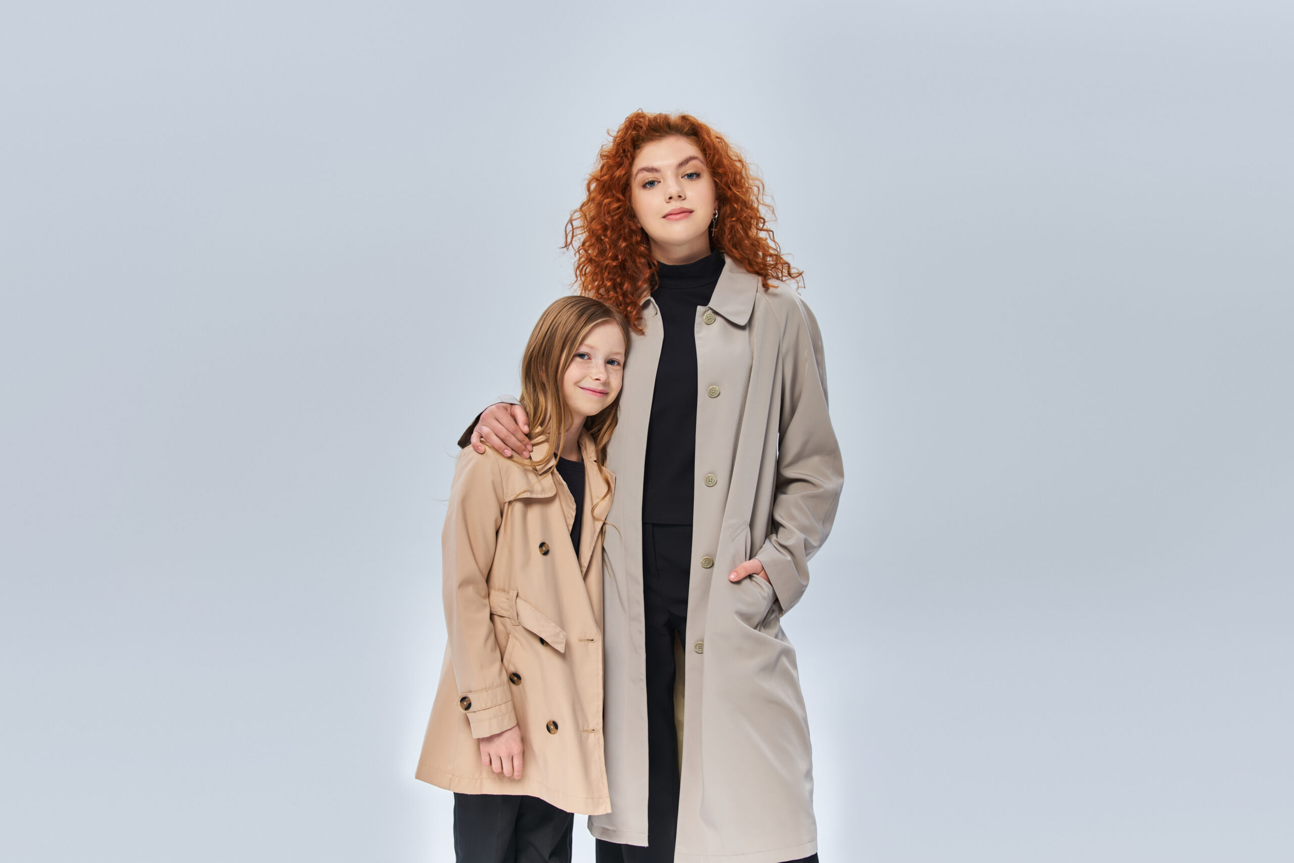 two female generations, redhead woman embracing daughter and standing in coats on grey background