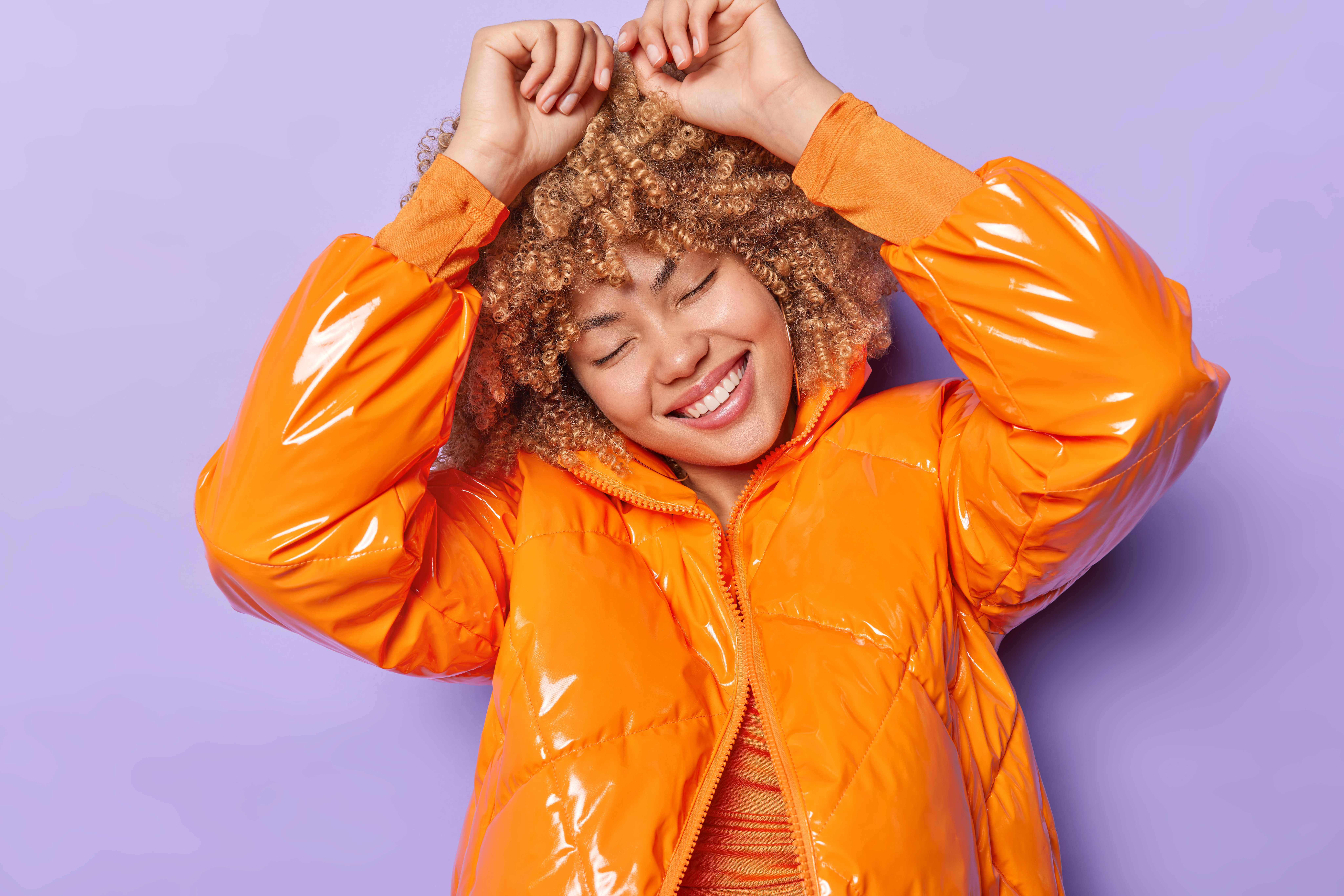 Waist up shot of carefree young woman with curly hair keeps arms raised up dressed in orange jacket keeps eyes closed smiles toothily has fun isolated over purple background enjoys happy moments