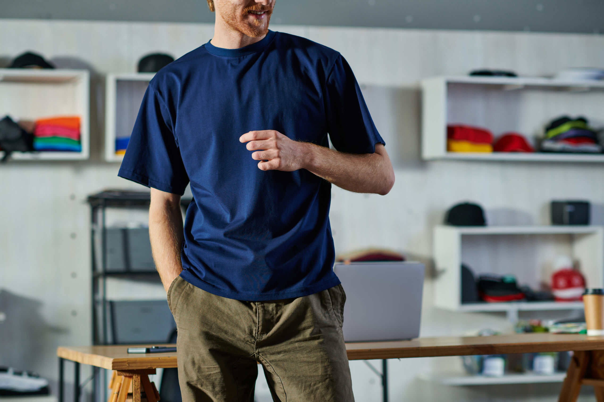 Cropped view of smiling young craftsman in casual clothes holding hand in pocket while standing near working table in print studio, self-made success concept